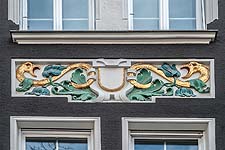 Farbiges Stuckornament mit goldenen Schlangen, Wappen? und floralen Elementen.